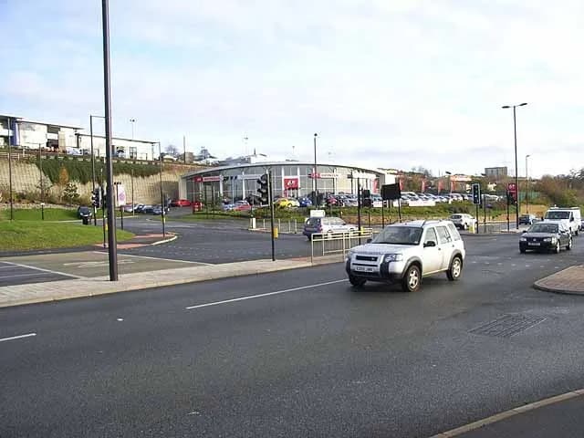 A silver SUV drives on a paved road at a complex urban intersection with a car dealership and other buildings in the background.