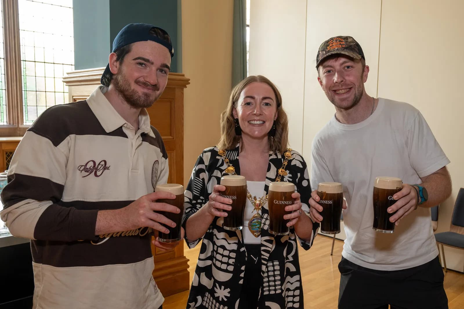 Three smiling people hold up glasses of Guinness beer, with the woman in the center wearing a ceremonial chain.