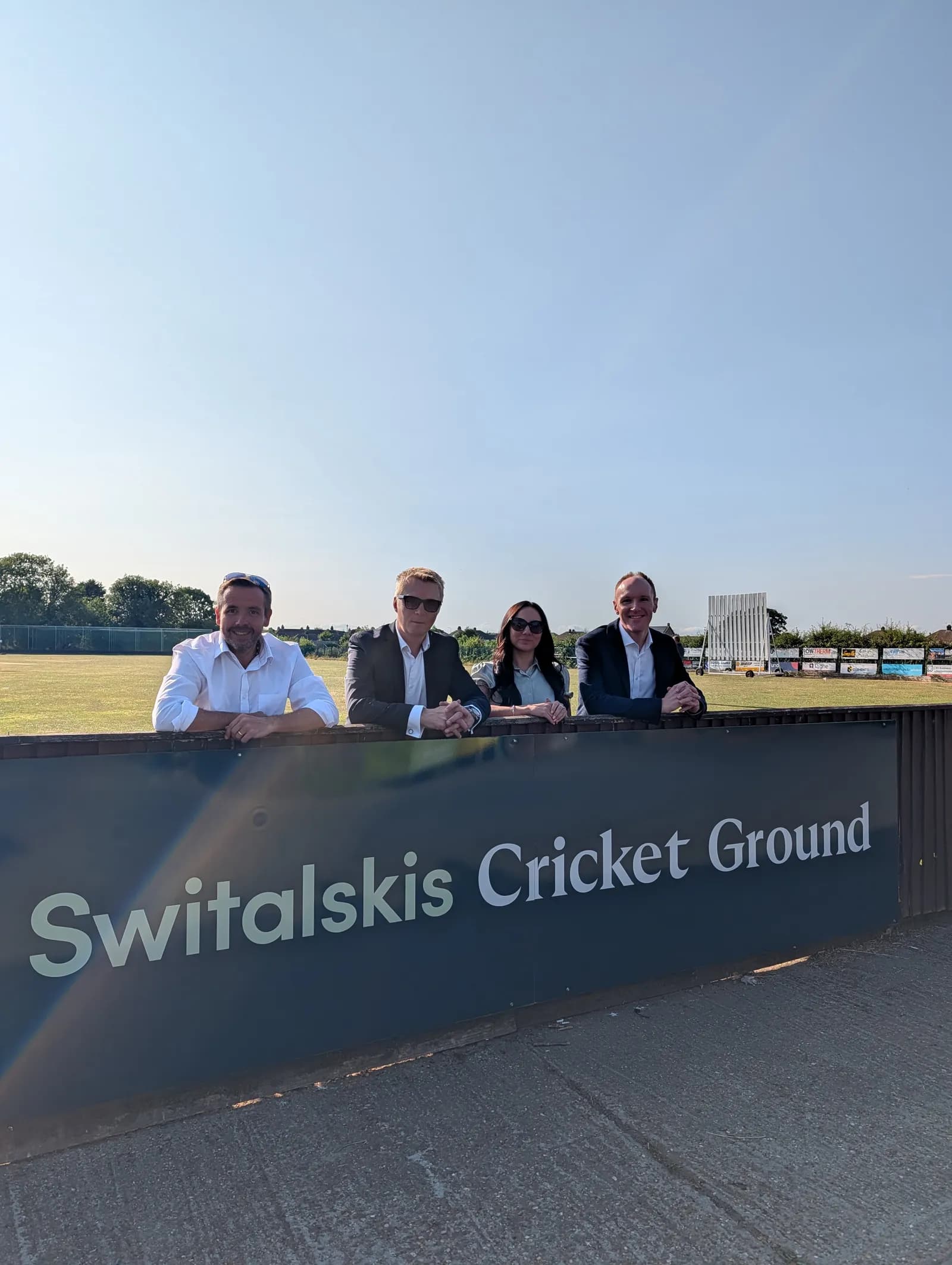 Four people, three men and one woman, are standing behind a sign that reads "Switalskis Cricket Ground" with a cricket field and clear blue sky in the background.
