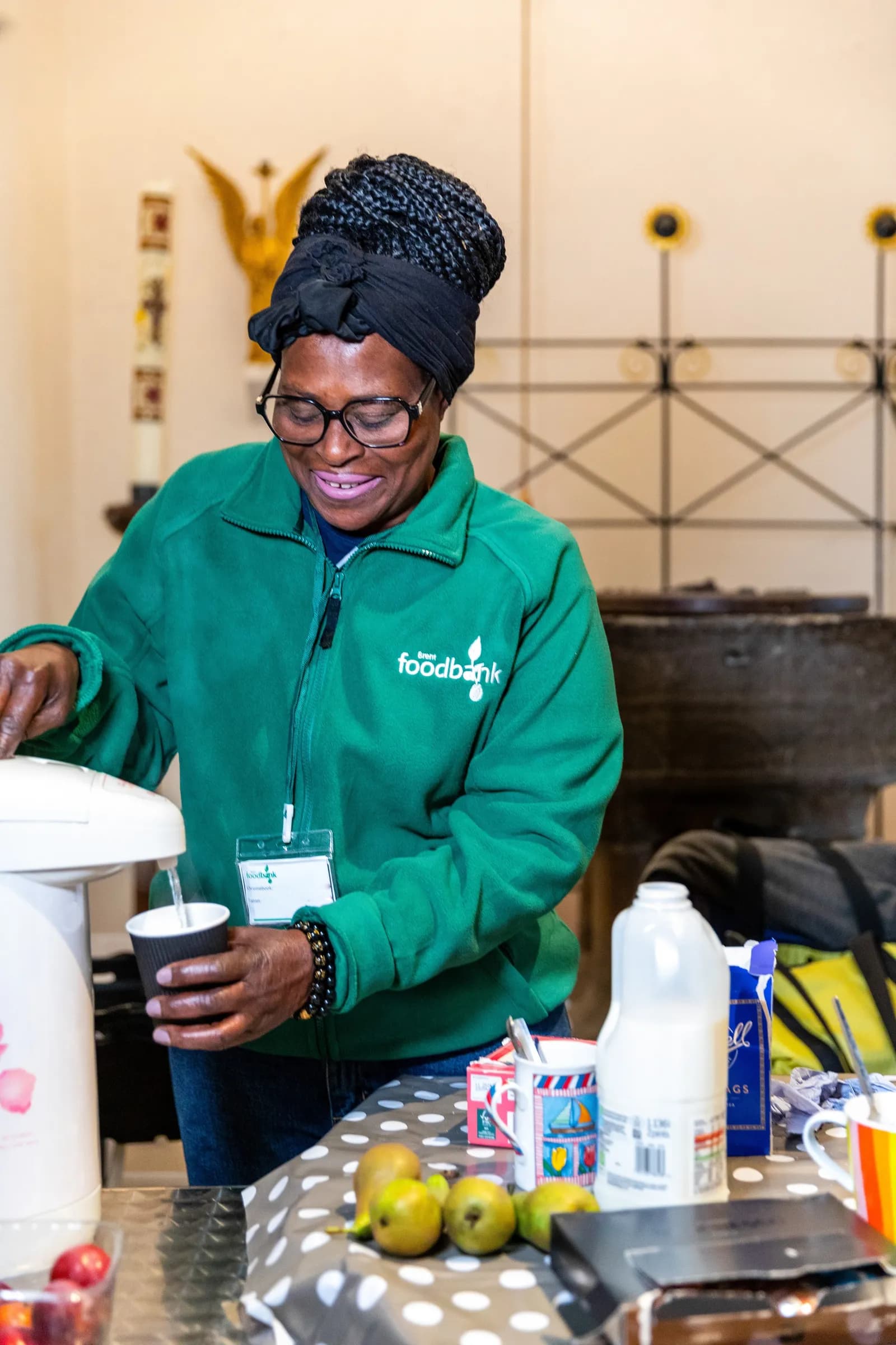 A woman wearing a green food bank jacket and a black head wrap smiles while pouring water into a cup from a dispenser on a table with fruit and milk.