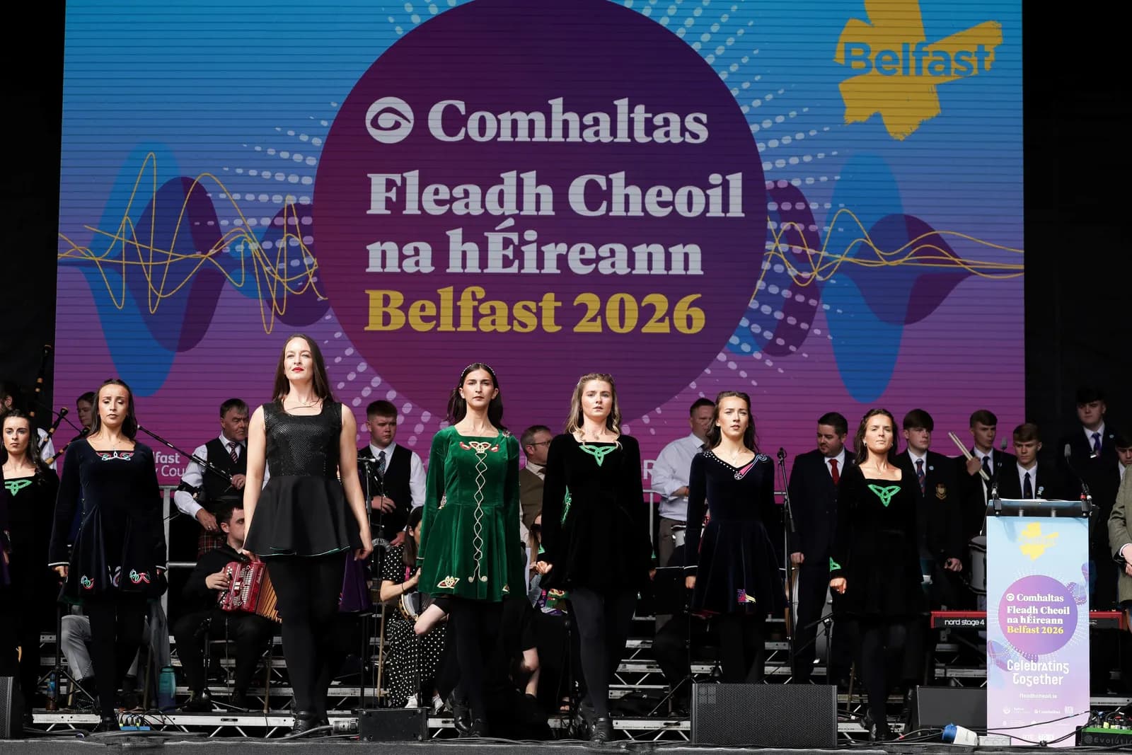 Several women in traditional Irish dance costumes stand on a stage in front of a large screen displaying the 'Comhaltas Fleadh Cheoil na hÉireann Belfast 2026' logo, with musicians and other performers in the background.