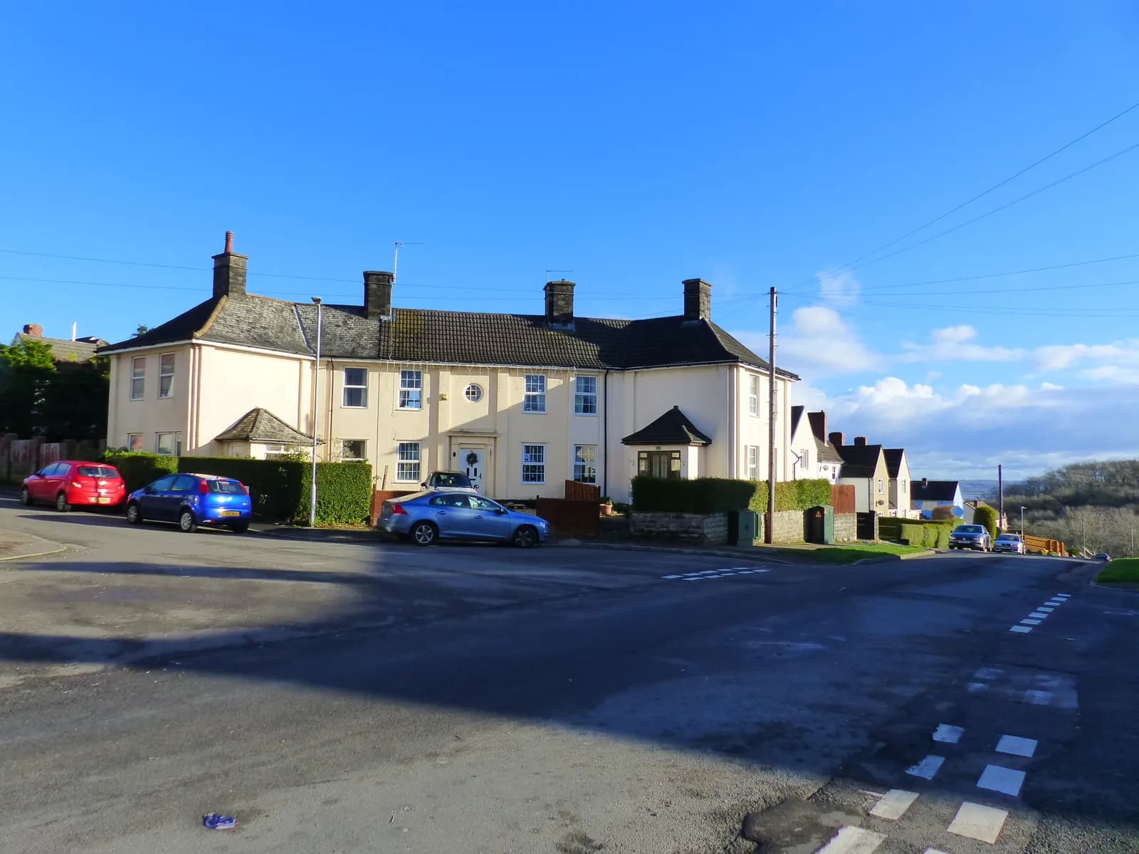 A row of light-colored houses with dark roofs line a street with cars parked along the side, under a clear blue sky.