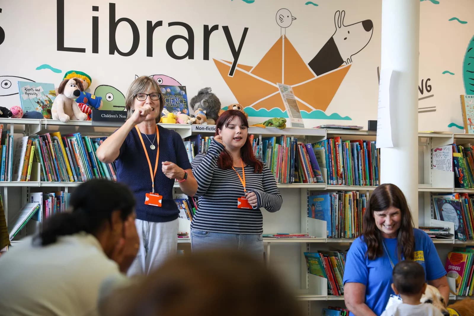 Two women, one older with glasses and one younger with red hair, stand in front of a bookshelf making hand gestures while a third woman smiles at a small child in the foreground.
