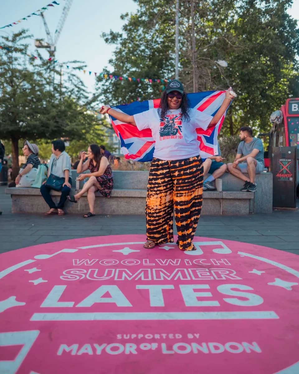 A woman stands in the center of a pink circular platform with text, holding a British flag open behind her, surrounded by people and trees.
