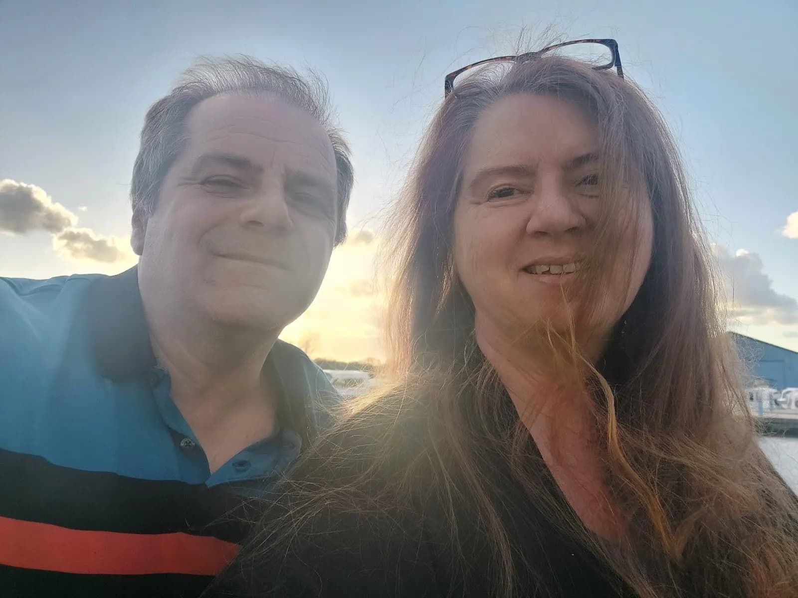 A man and a woman pose closely together for a selfie outdoors under a bright sky with sun flare.