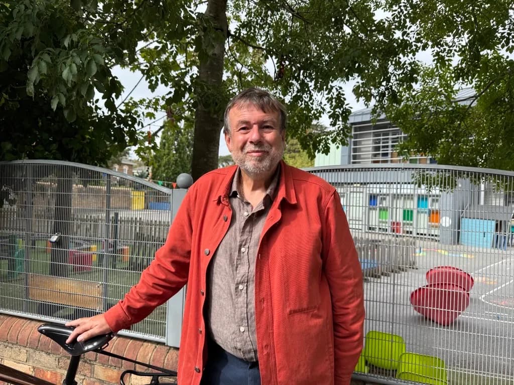 A man with a beard and an orange jacket stands next to a bicycle with a school playground in the background.