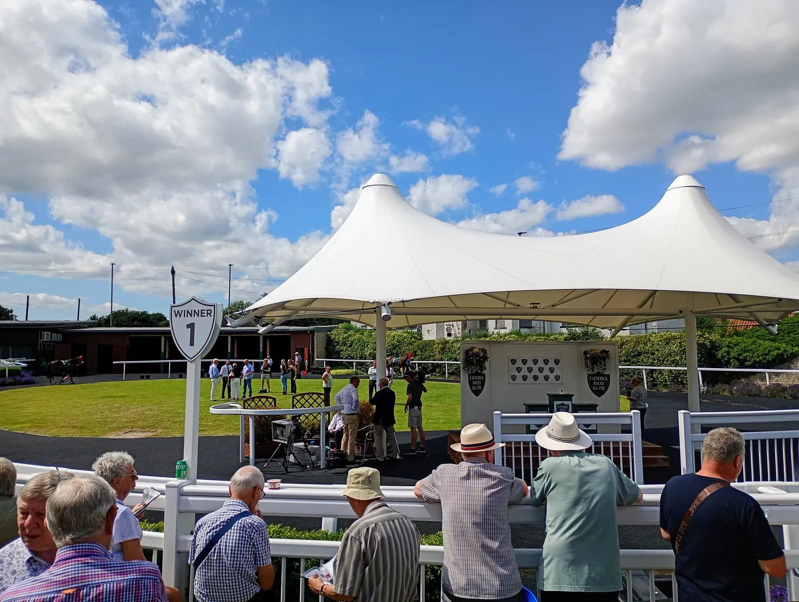 A crowd of people at a horse racing event under a large white tent, with a "Winner 1" sign in the foreground.
