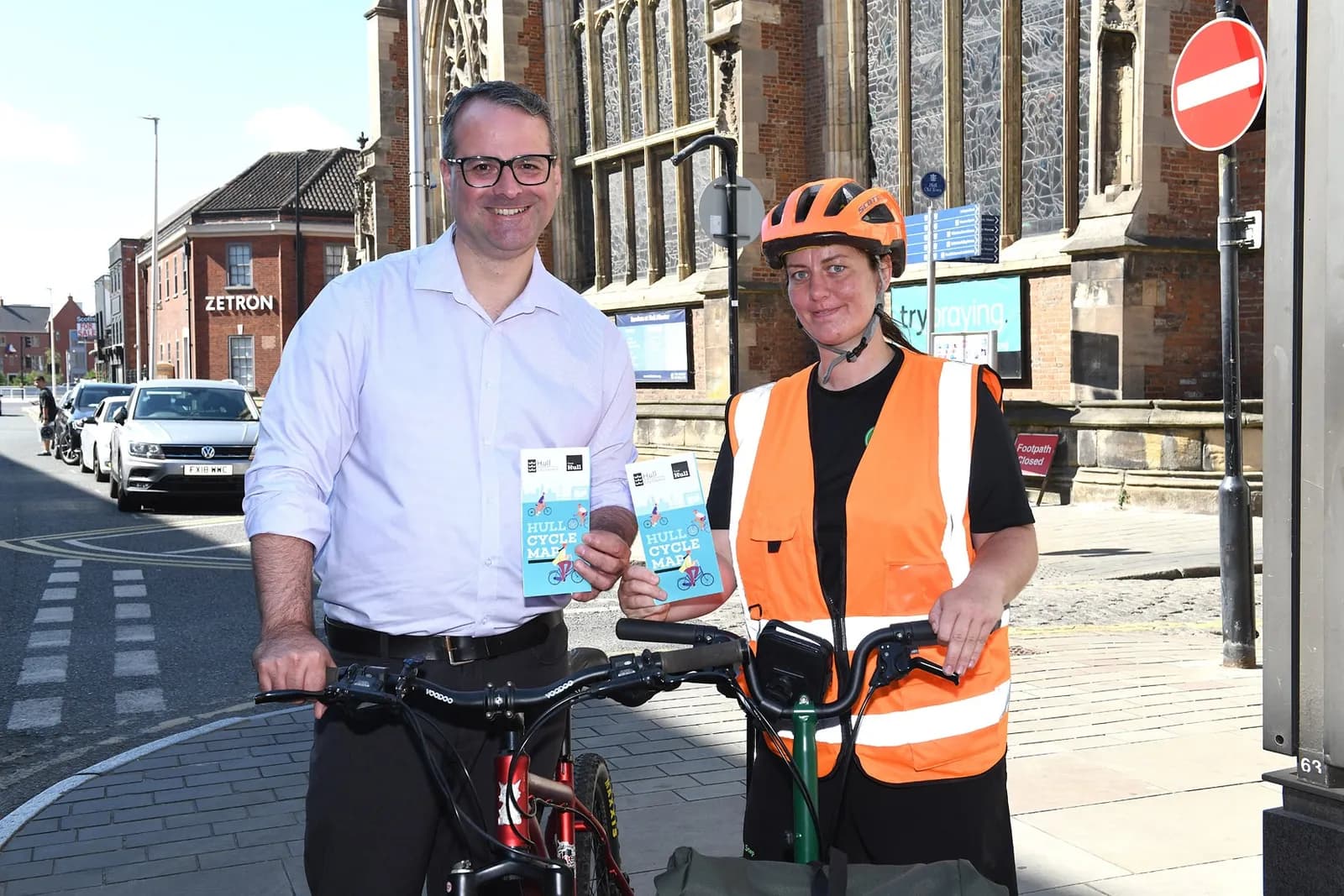 A man in a light purple shirt and glasses stands next to a woman in an orange safety vest and bike helmet, both holding "Hull Cycle Map" brochures and standing with bicycles on a city street.