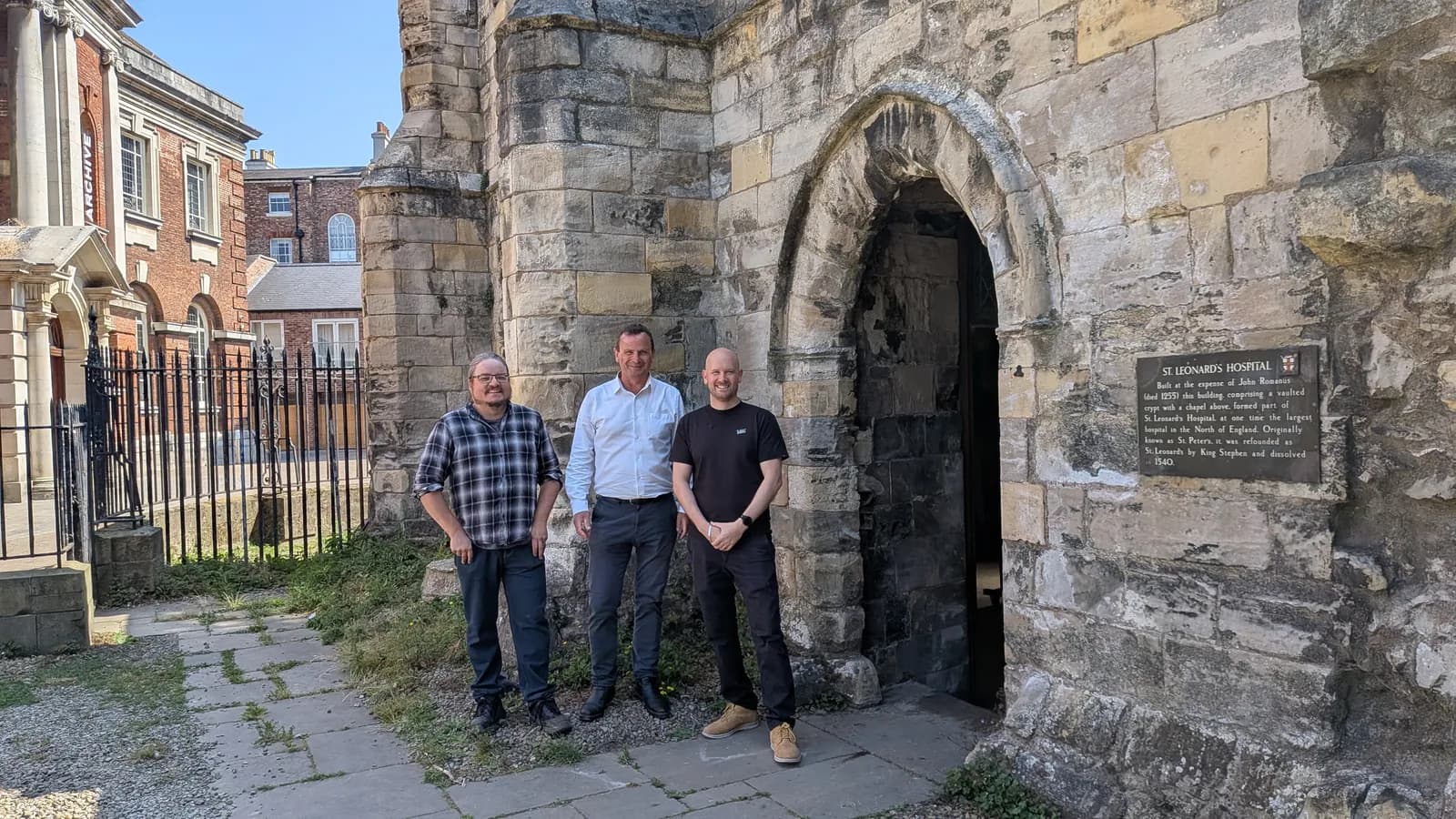 Three men stand smiling in front of a stone building with an arched entrance and a historical plaque, with another brick building visible in the background.