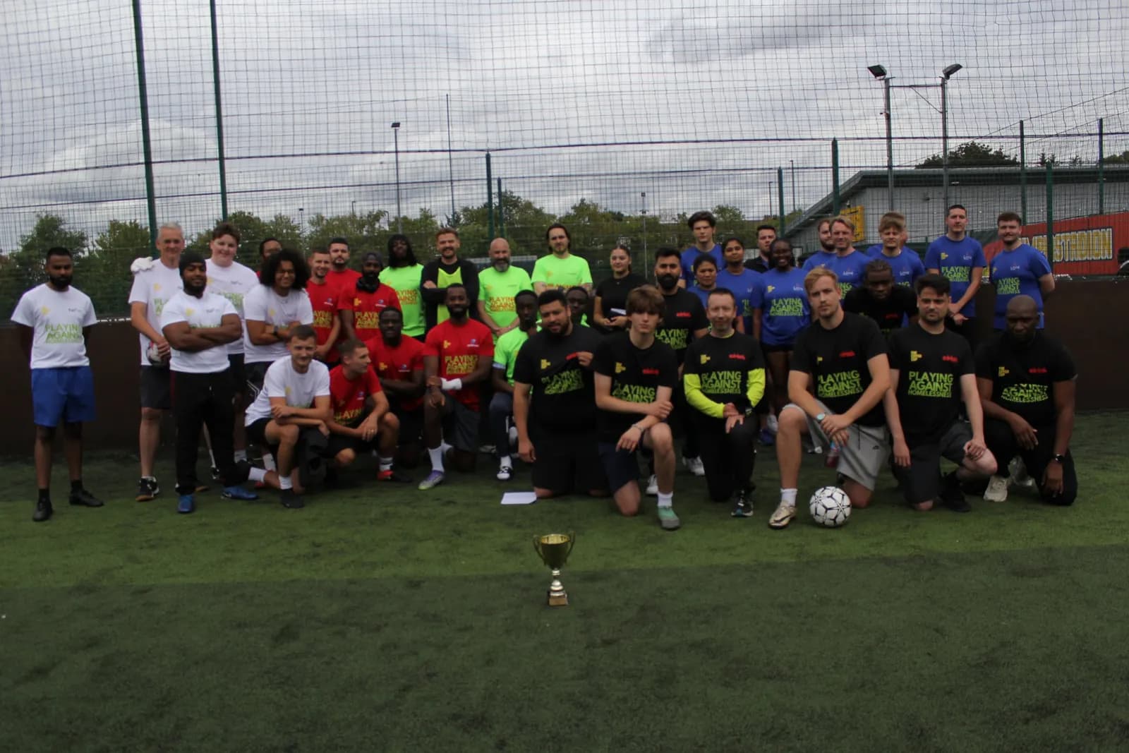 A large group of diverse people in various colored t-shirts and shorts stand and kneel on an artificial turf field, with a small golden trophy and a soccer ball in the foreground.