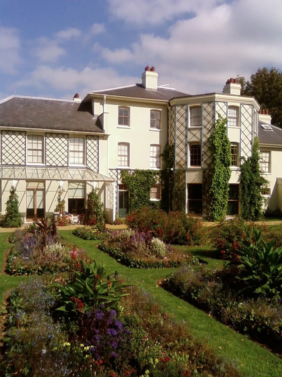 A large cream-colored house with a dark roof and decorative trellises on its facade overlooks a vibrant, meticulously landscaped garden with various flowering plants and green lawns under a blue sky.