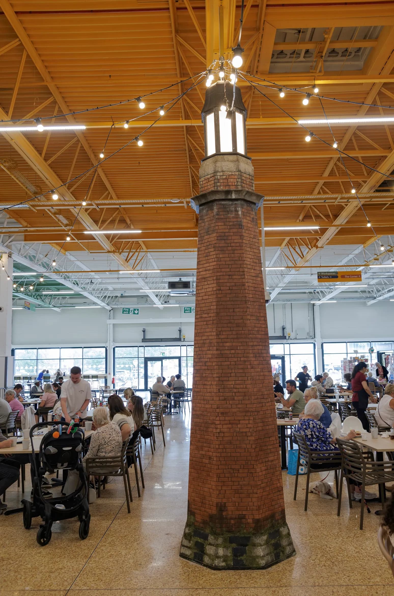 A tall, brick lighthouse replica stands prominently in the center of a bustling cafeteria with many people seated at tables, surrounded by bright overhead lighting and a high yellow ceiling.