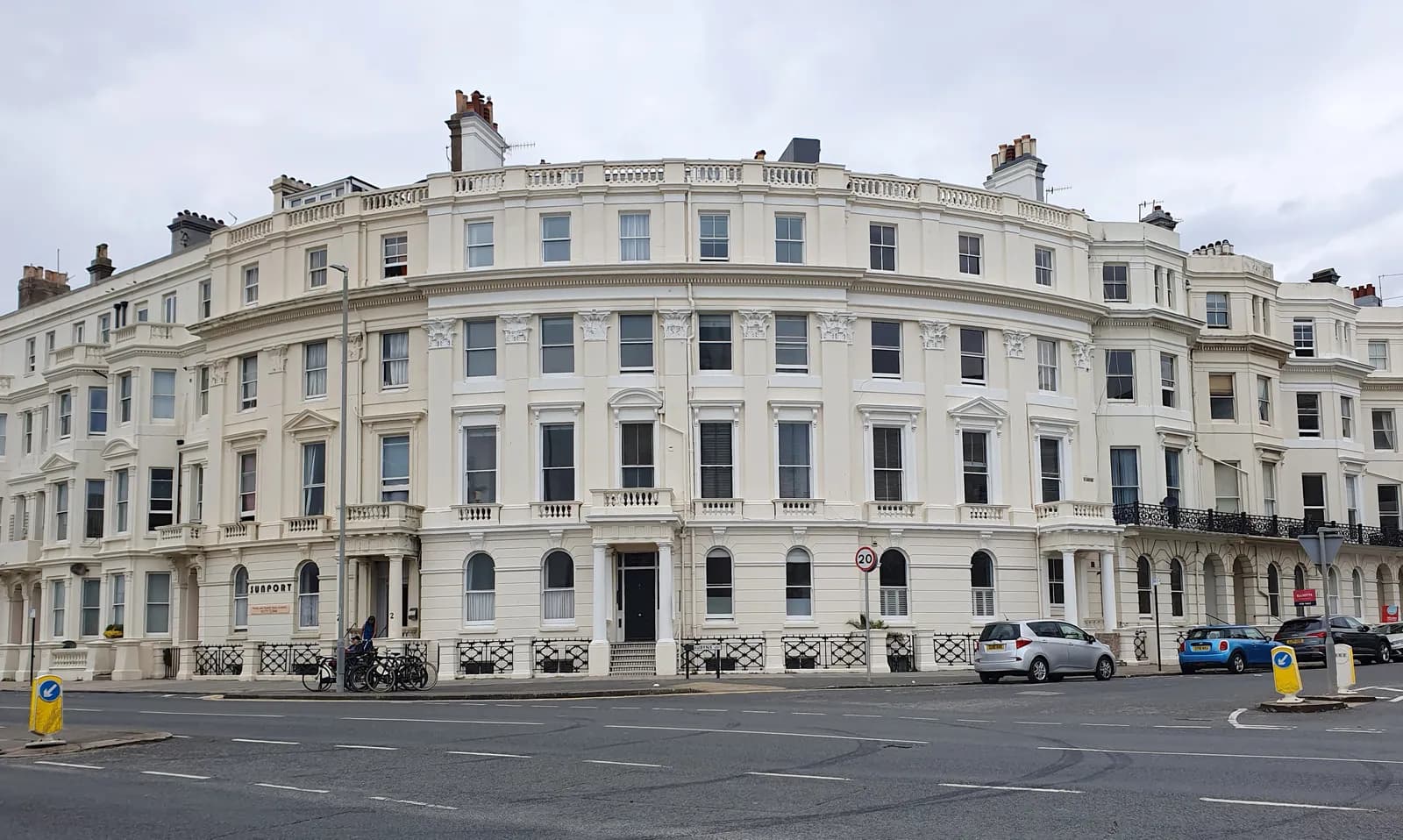 A large, curved white building with many windows and intricate architectural details stands beside a paved road with cars parked along the curb.