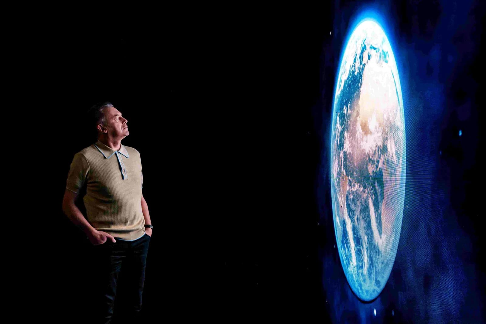 A man in a beige polo shirt looks up at a glowing, vertically oriented image of Earth against a dark background.