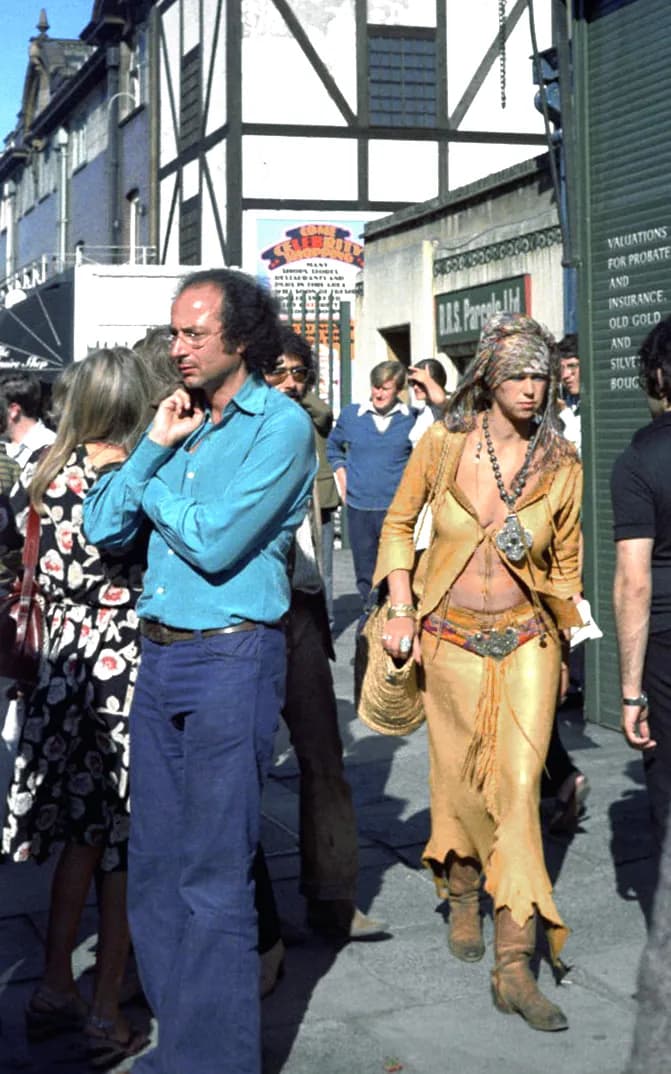 A man in a blue shirt and jeans with curly brown hair stands on a street with a woman in a floral dress on his left and a woman in a long bohemian outfit on his right.