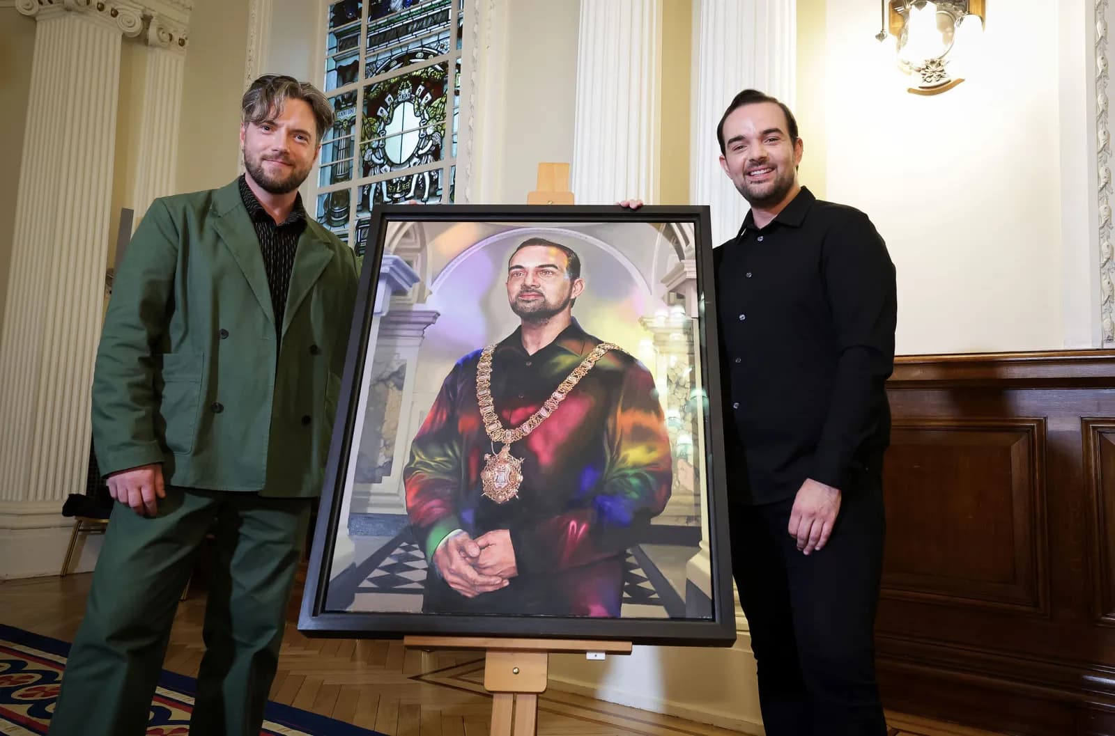 Two men standing on either side of a painted portrait, with the man on the left wearing a green suit and the man on the right wearing a black shirt, both smiling at the camera.