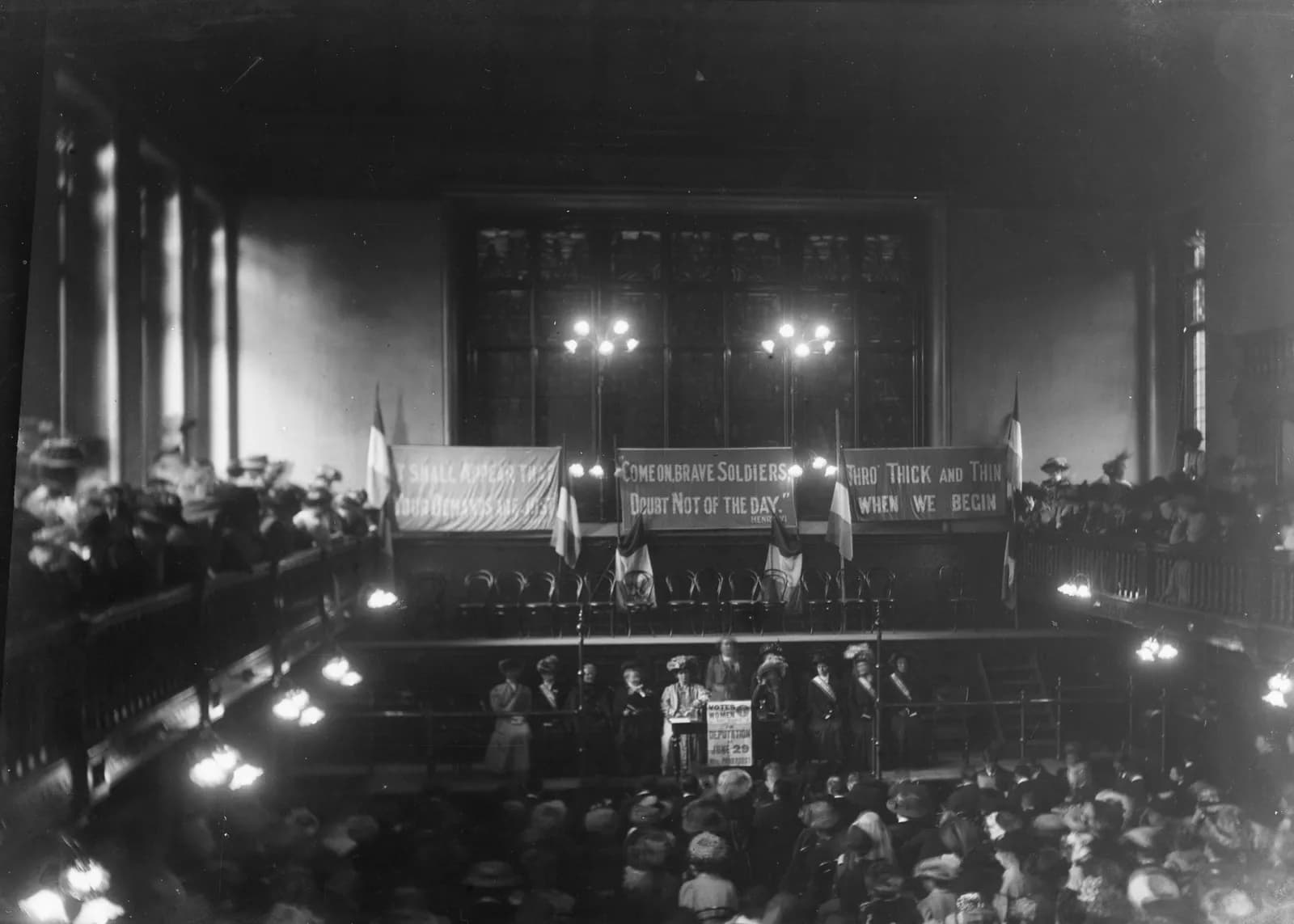 A public indoor gathering of mostly women with banners on the wall behind a speaker on a stage.