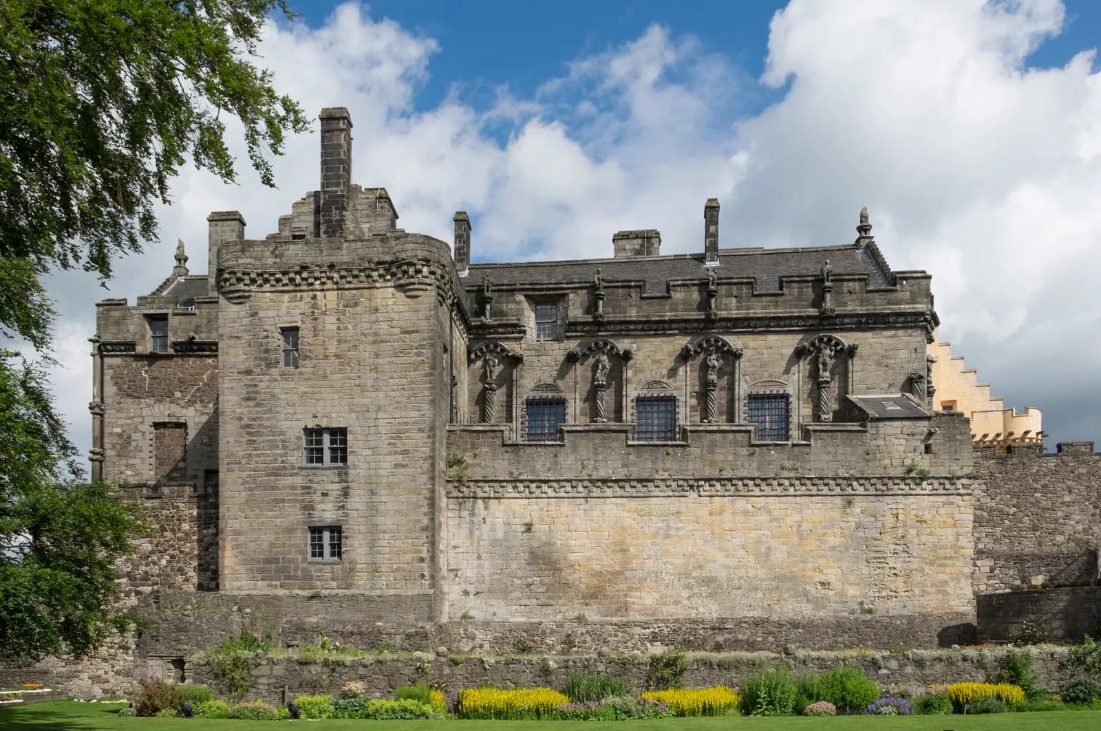A historic stone castle with multiple windows and ornate carvings stands against a cloudy blue sky, with a green lawn and flowerbeds in the foreground and a tree branch partially obscuring the left side of the building.