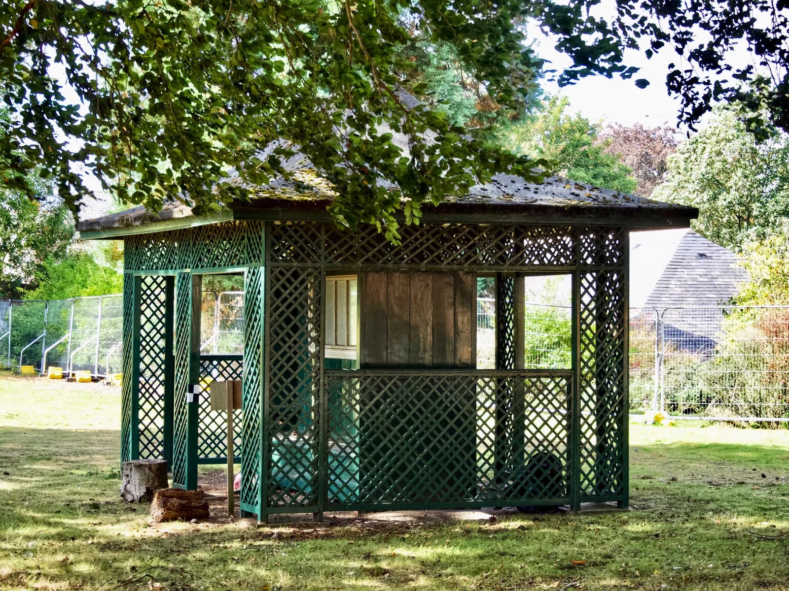 A green wooden gazebo with a lattice pattern stands on a grassy lawn under overhanging tree branches.