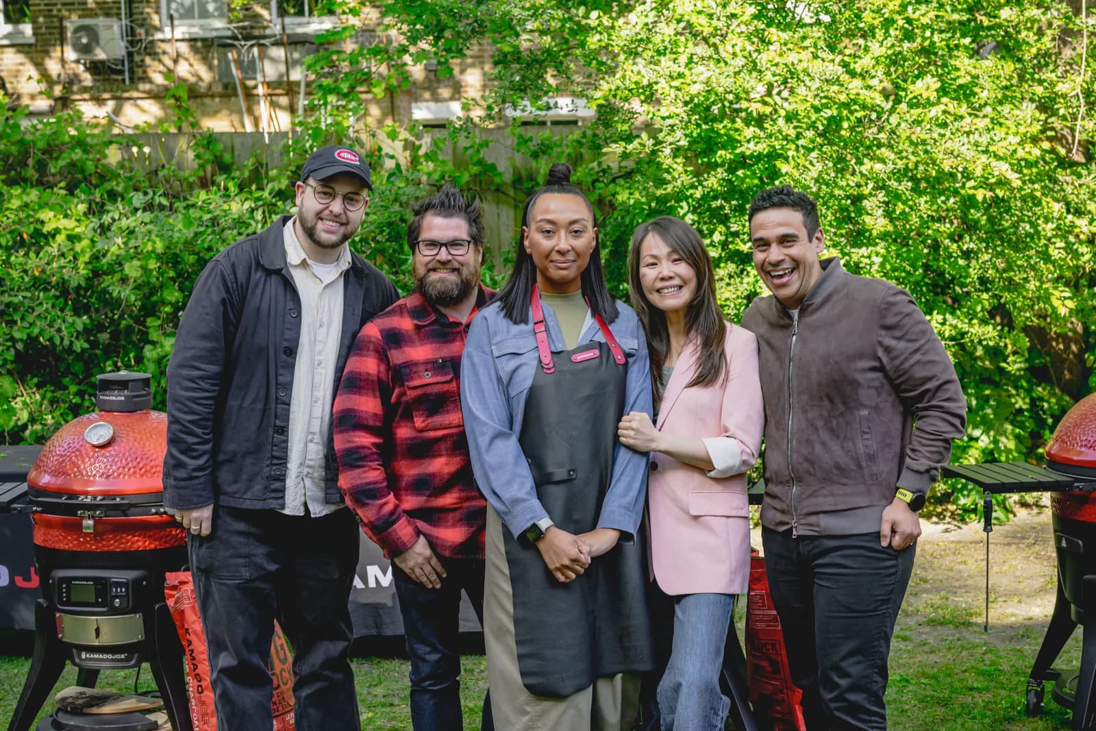 Outdoor portrait of five smiling people, three men and two women, dressed casually and standing before lush green foliage, with a red grill visible on the left.