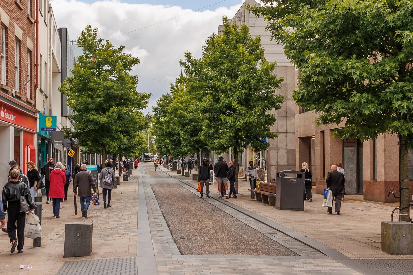 A wide pedestrianized street lined with shops and trees, with numerous people walking along the sidewalks and a few in the central pathway.