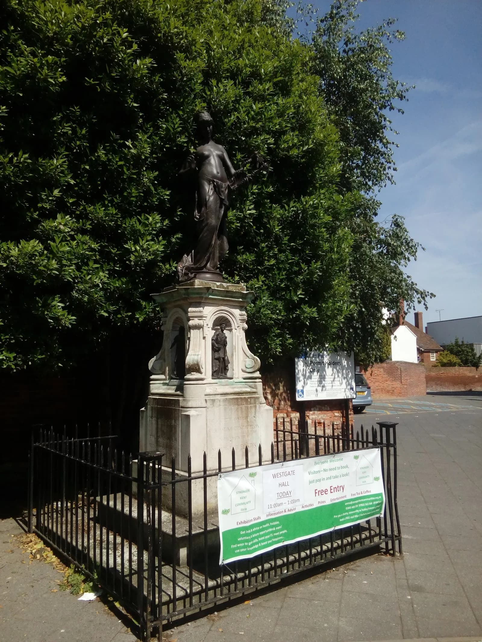 A bronze statue of a female figure holding a wreath, standing on a stone pedestal surrounded by a black metal fence, with green trees in the background.
