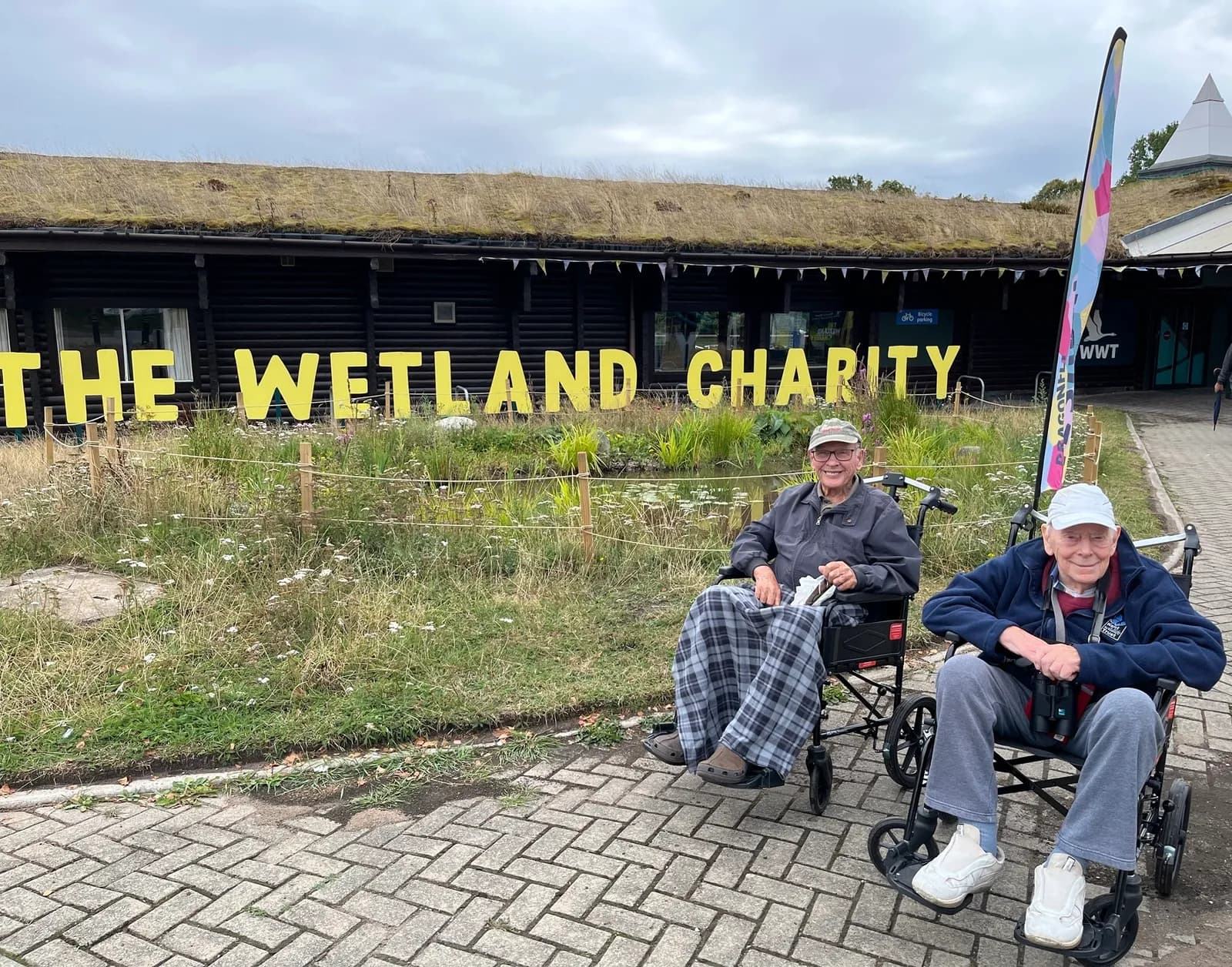 Two elderly men in wheelchairs smiling in front of a building with a green roof and large yellow letters spelling "THE WETLAND CHARITY".
