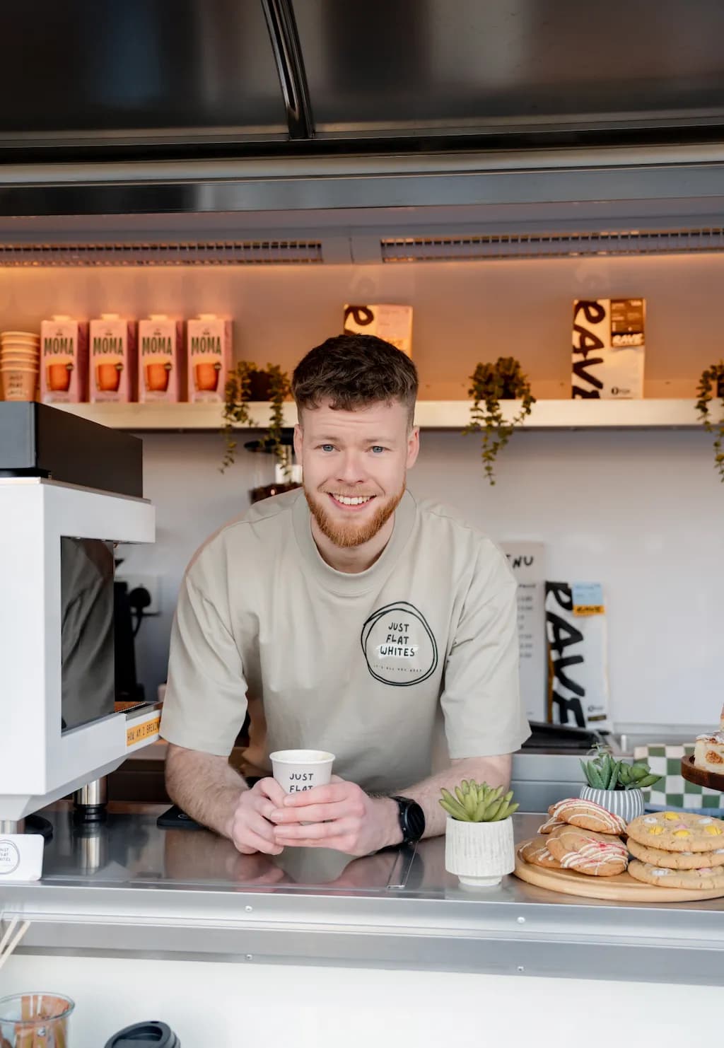 A smiling young man with a beard and short hair, wearing a light brown t-shirt with a 'Just Flat Whites' logo, stands behind a counter in what appears to be a mobile coffee shop or food truck, holding a coffee cup in his hands.