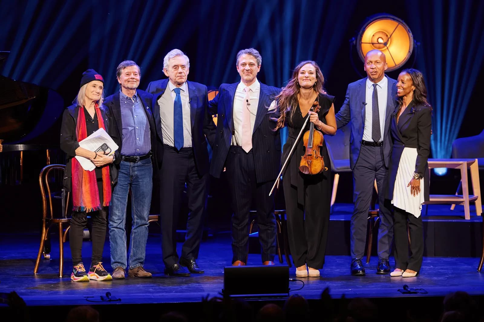 Seven people, including two women and five men, stand on a stage with a violin, mic, and piano in the background.