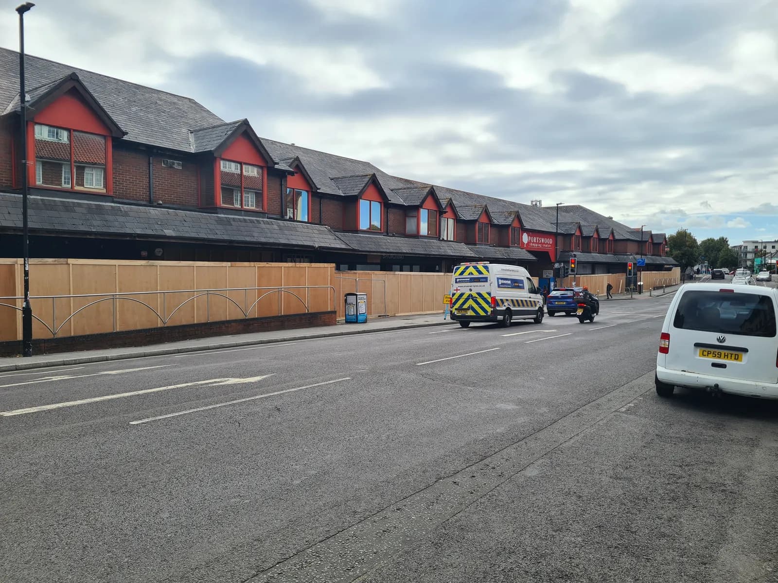A long, red-roofed building with several dormer windows lines a street with parked and moving vehicles, all under a cloudy sky.