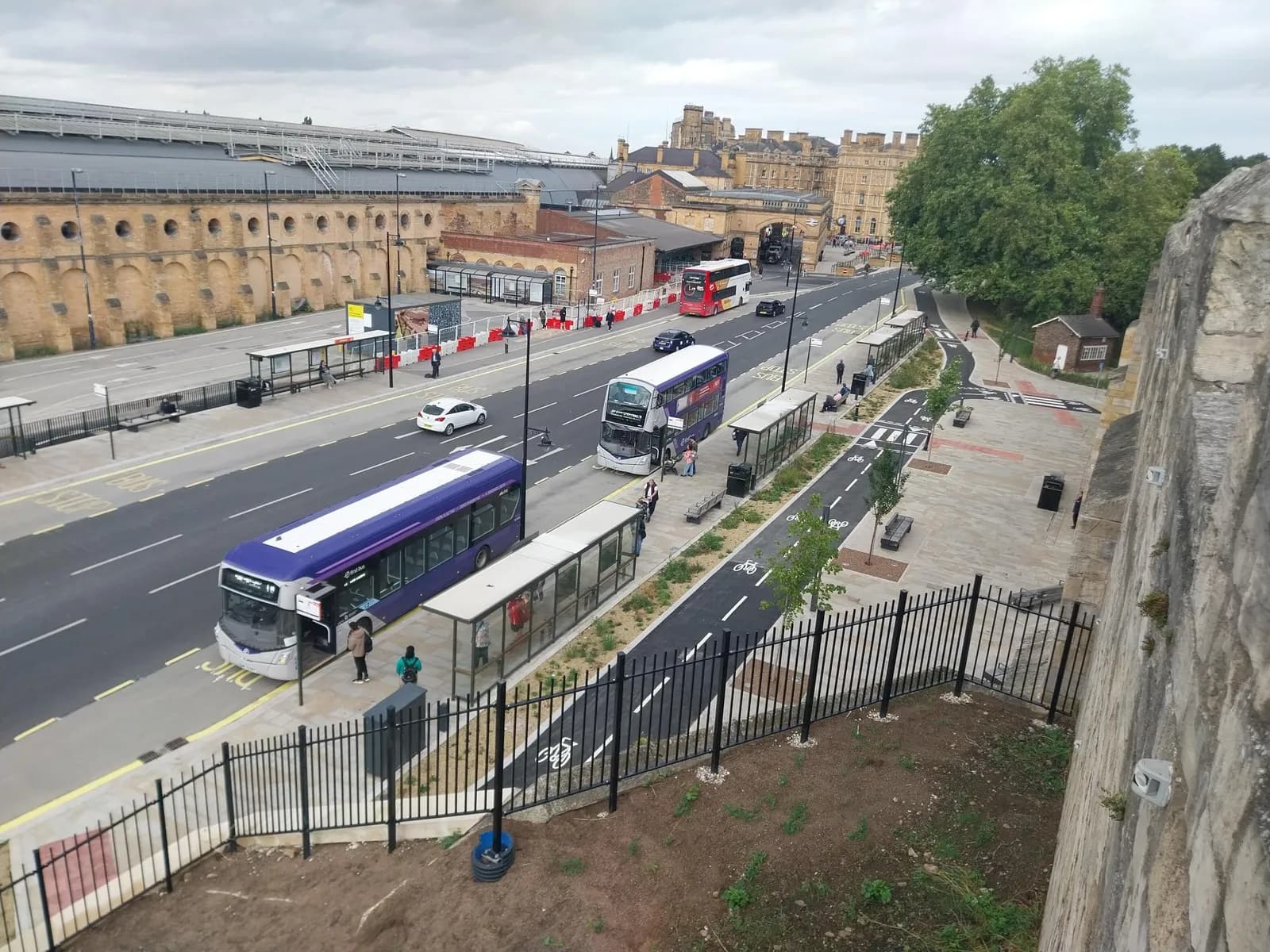 An overhead view of a street with multiple buses, cars, bus stops, and a bike lane, all set against a backdrop of historic buildings and a large tree.