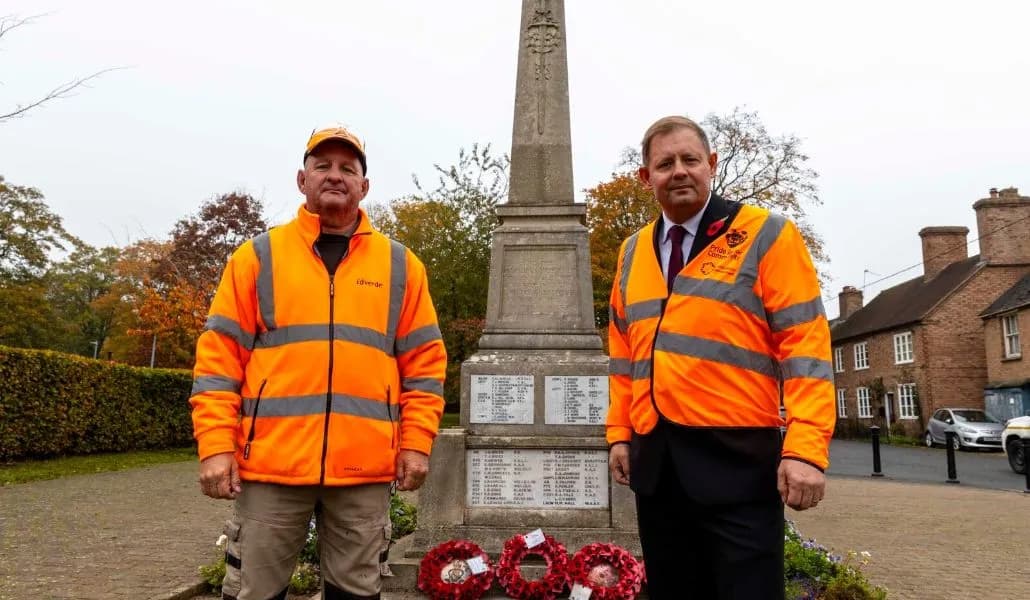 Two men in orange high-visibility vests stand solemnly on either side of a war memorial adorned with poppy wreaths.