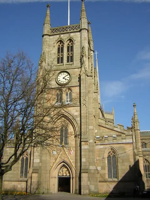 A tall, ornate stone church tower with a clock and arched windows stands against a clear blue sky, with a bare tree partially obscuring the left side of the building.