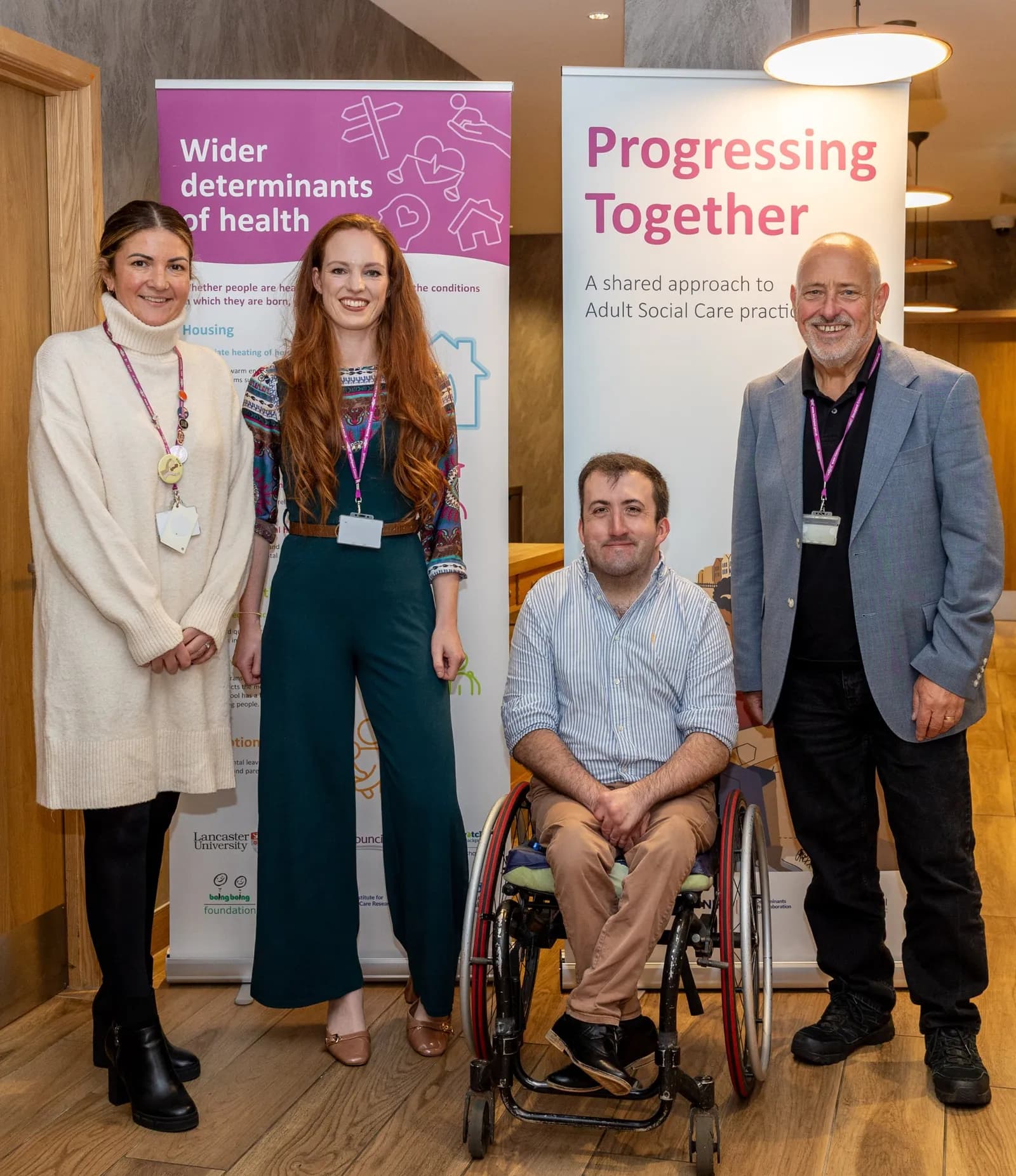 Four people, two men and two women, stand or sit in front of two banners. One woman is on the far left, wearing a cream-colored sweater and black boots. Next to her is a woman with long red hair wearing a patterned top and dark green jumpsuit. A man in a blue and white striped shirt sits in a wheelc