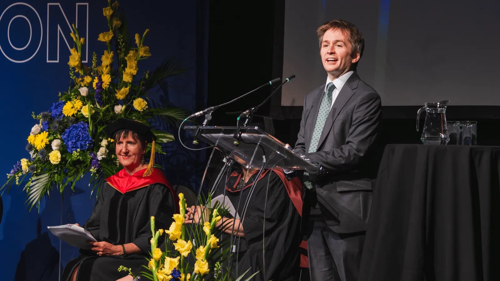 A man in a suit speaks at a podium, while a woman in graduation attire sits nearby, with floral arrangements on a stage with a blue backdrop.