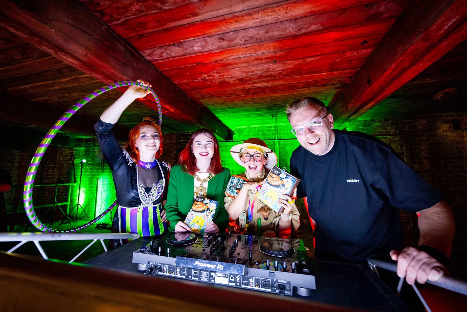 Four people, including one DJ, smiling and posing with an album behind a DJ deck, illuminated by green and red lighting from above.