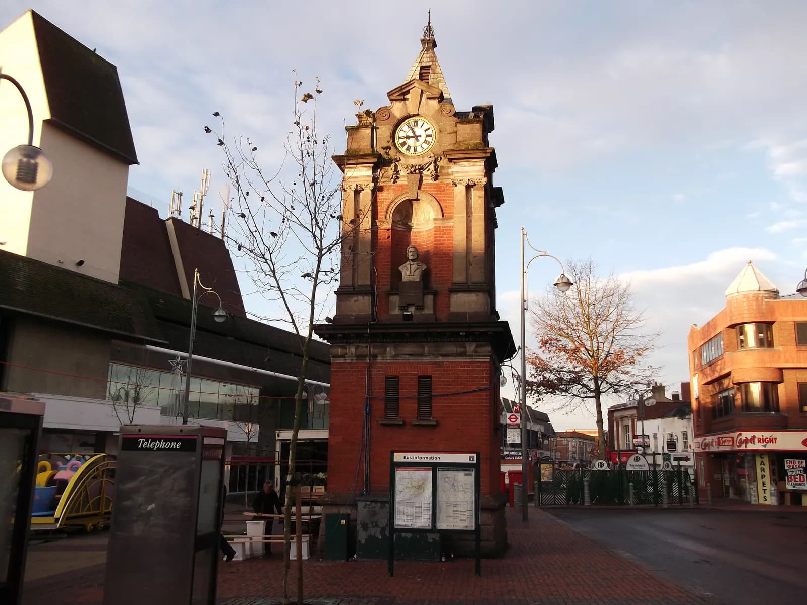 A red brick clock tower topped with a bust statue stands prominently on a street corner, with other buildings and a bare tree under a cloudy sky.