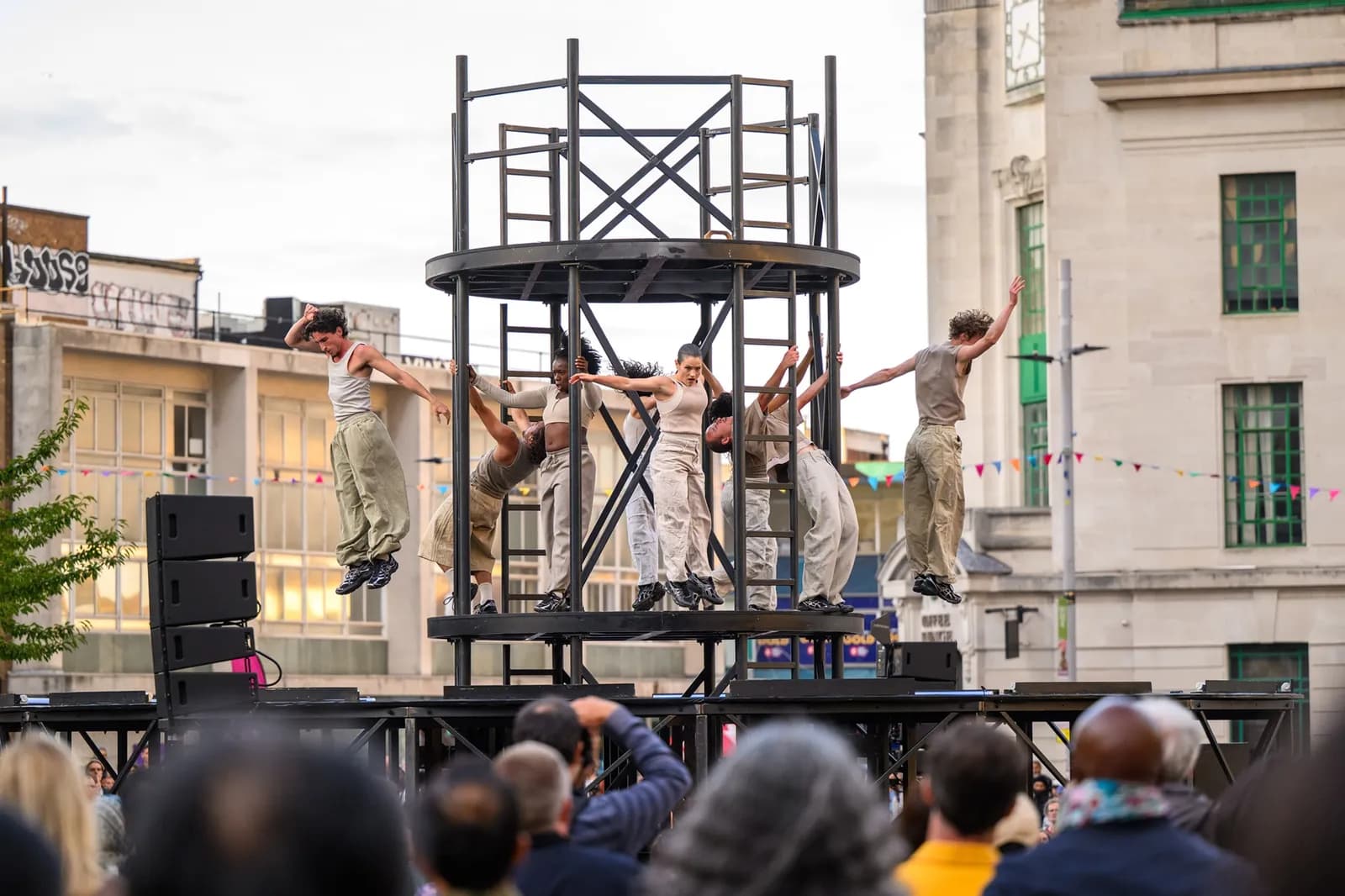 A group of dancers in tan outfits perform on a multi-level stage in a city street, with one dancer suspended in mid-air.