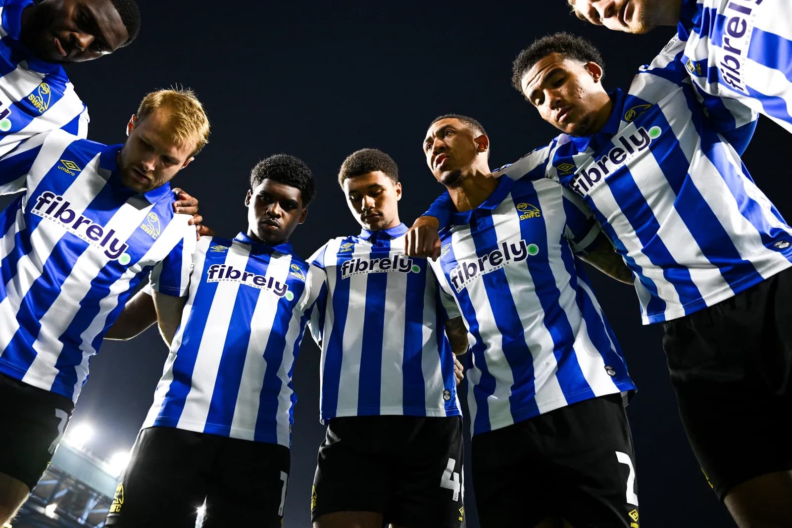 A group of six male soccer players in blue and white striped jerseys, seen from below, huddle together with their arms around each other's shoulders.