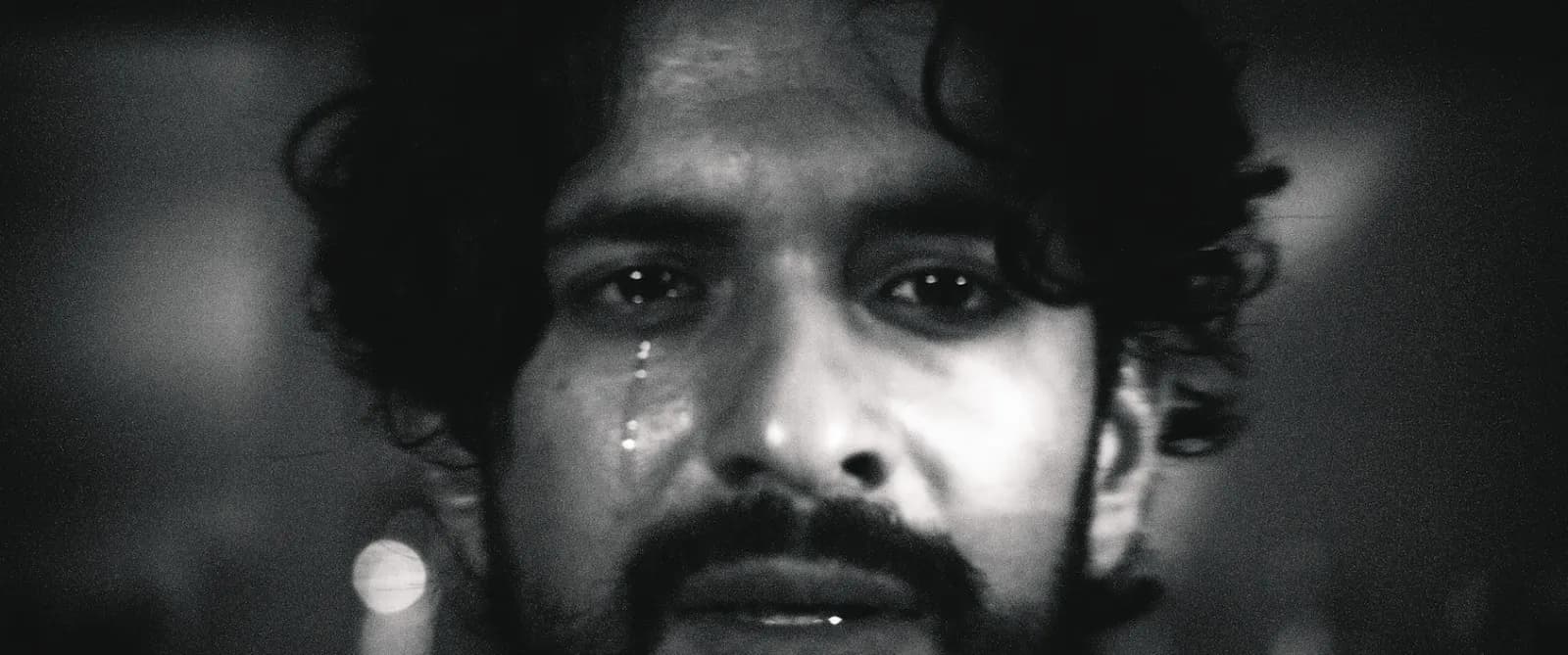 A distressed man with curly hair and a beard, in black and white, has tears streaming down his face.