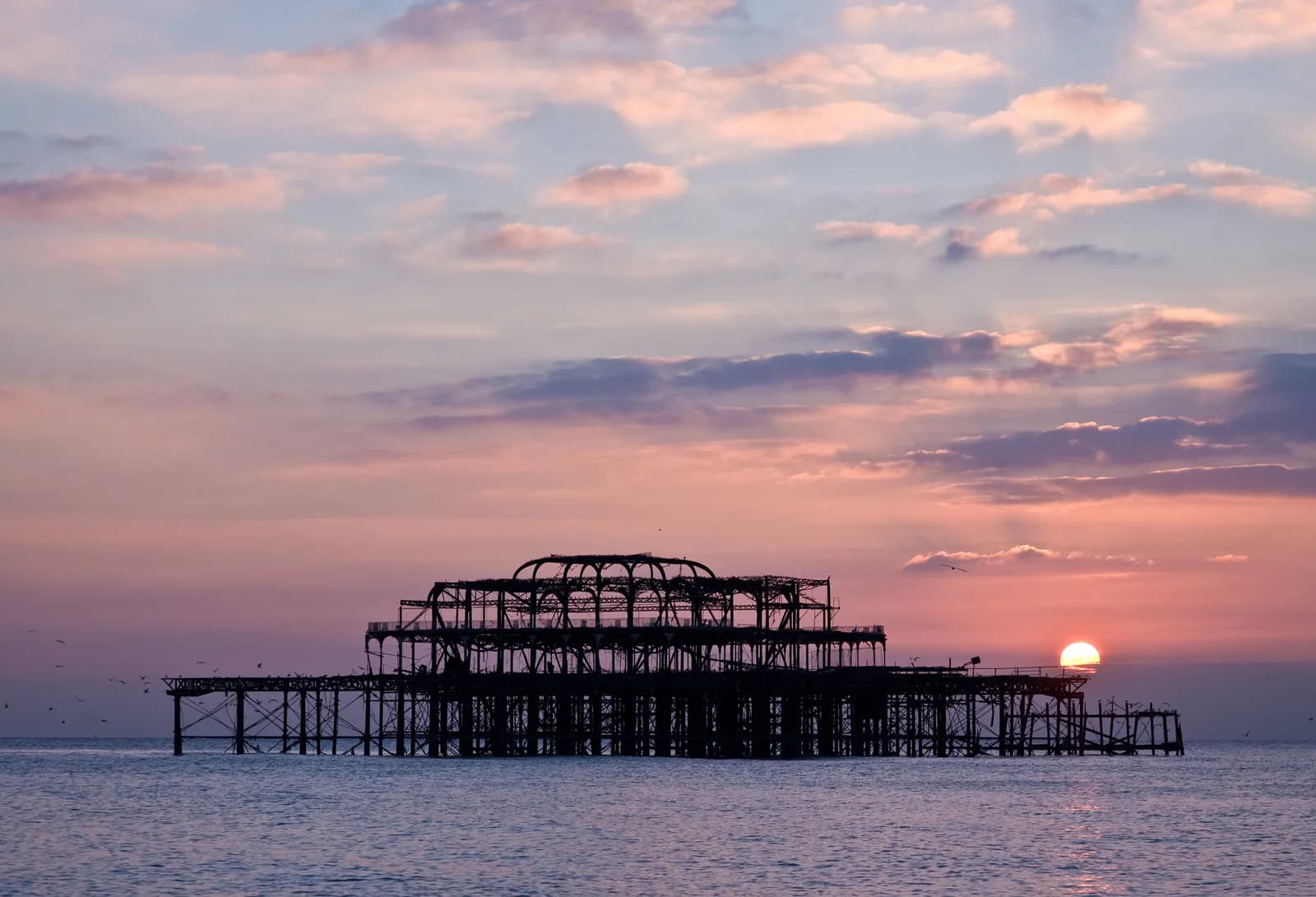 A silhouette of a derelict Brighton West Pier in the sea at sunset, with birds flying around and a visible pink and blue sky.