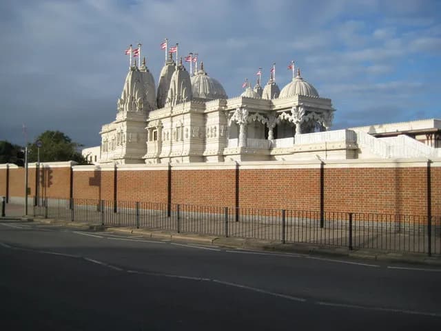 A large, ornate white temple with multiple domed and pointed towers, adorned with small flags, stands behind a brick wall bordering a street.