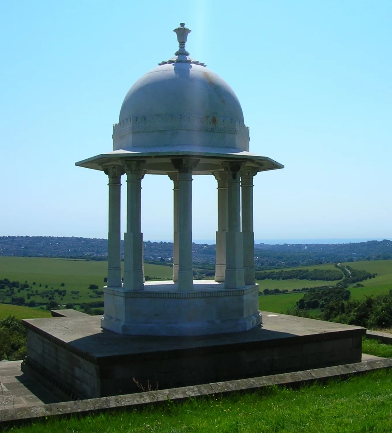 A white domed monument with columns stands on a concrete base overlooking a vast green landscape with a distant town and ocean under a clear blue sky.