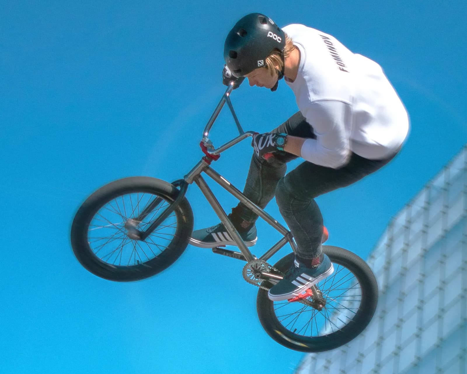 A BMX rider in mid-air, wearing a black helmet, white long-sleeve shirt, and dark pants, performing a trick against a bright blue sky with a building partially visible in the background.