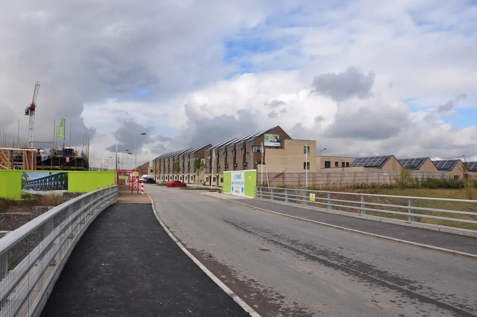 A long paved road leads towards a row of modern terraced houses with solar panels on their roofs, with construction on the left and a grassy area with wooden fences on the right, under a cloudy sky.