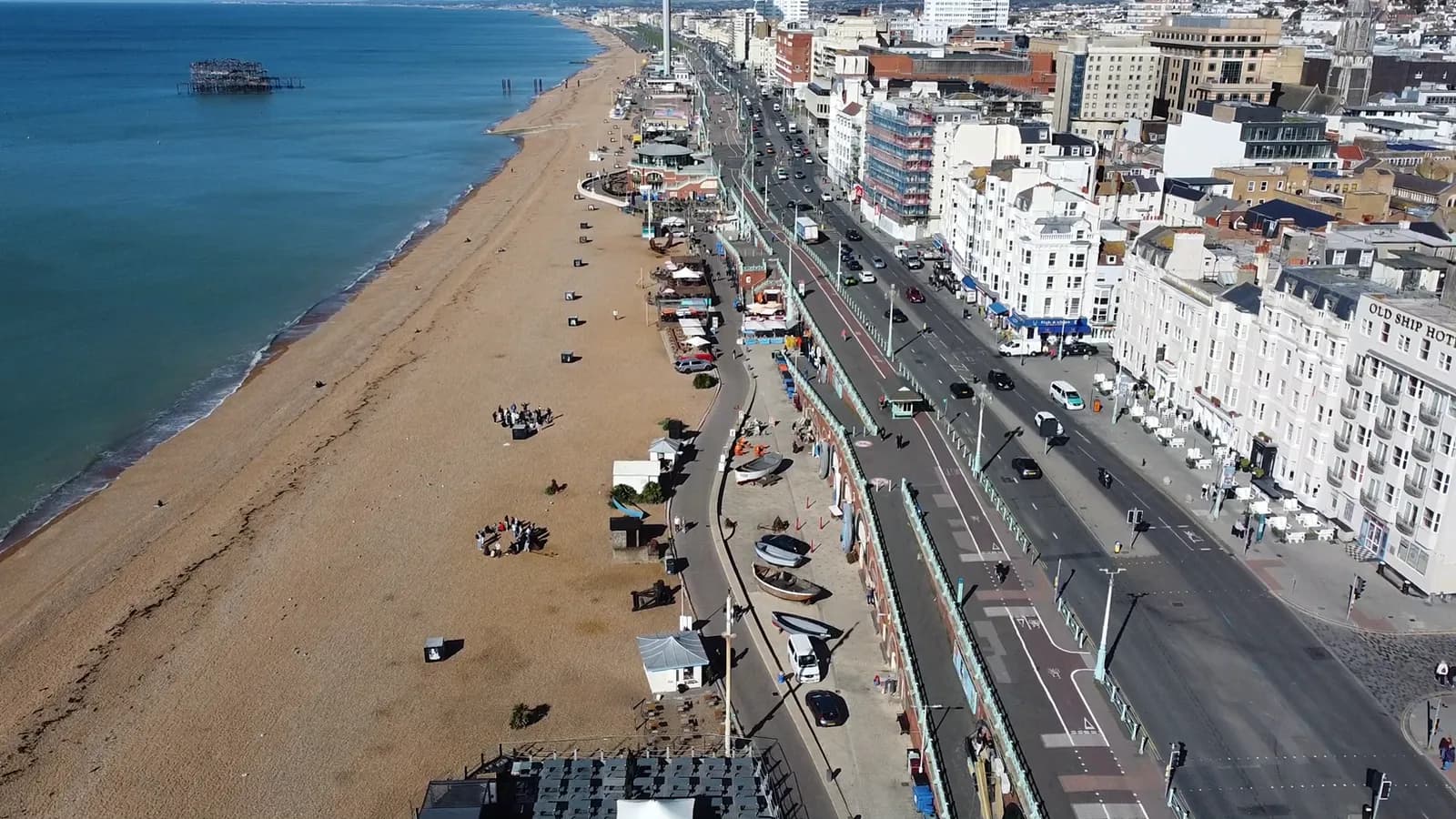 An aerial view captures the vibrant Brighton seafront, featuring a wide sandy beach, the serene blue sea with the distant West Pier, and a bustling coastal road lined with buildings and vehicles.
