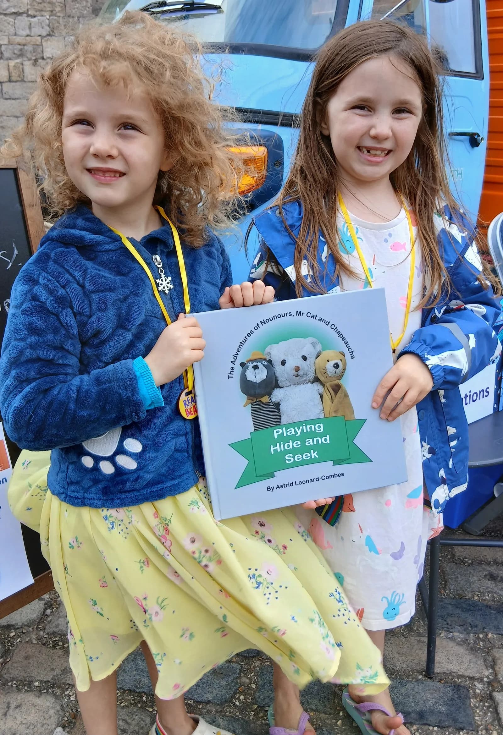 Two young girls are smiling while holding a children's book titled "Playing Hide and Seek", featuring three teddy bears on the cover.