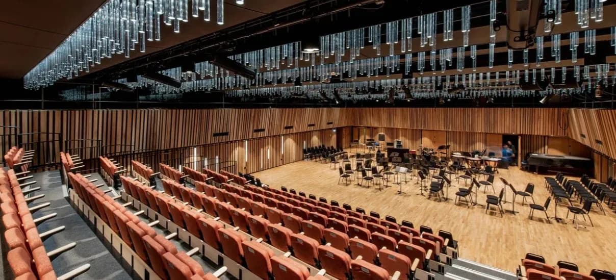 An empty modern concert hall with rows of orange seats in the foreground and a wooden stage with music stands and chairs in the background, illuminated by hanging cylindrical lights.