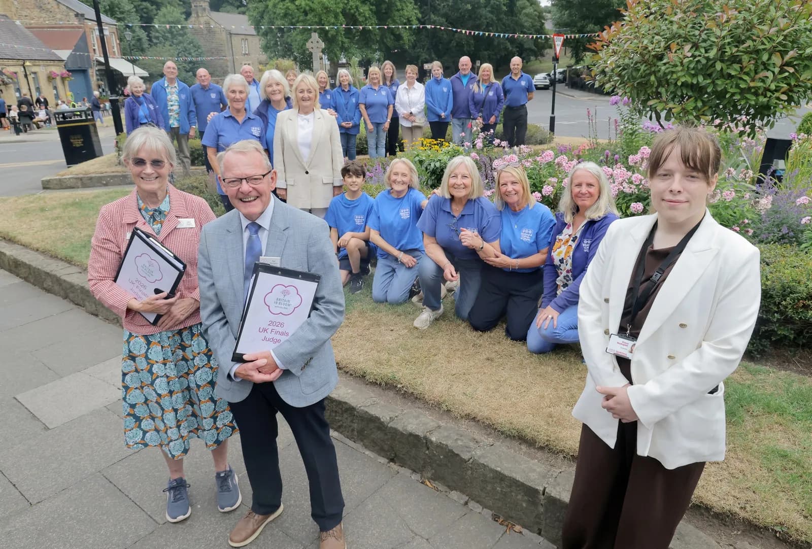 A large group of people of varying ages stand and kneel in front of a flowerbed and buildings, many wearing blue shirts, with two individuals in the foreground holding certificates.
