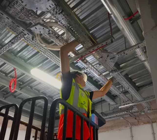 A construction worker wearing a hard hat and safety vest installs electrical conduits and cables on a ceiling.